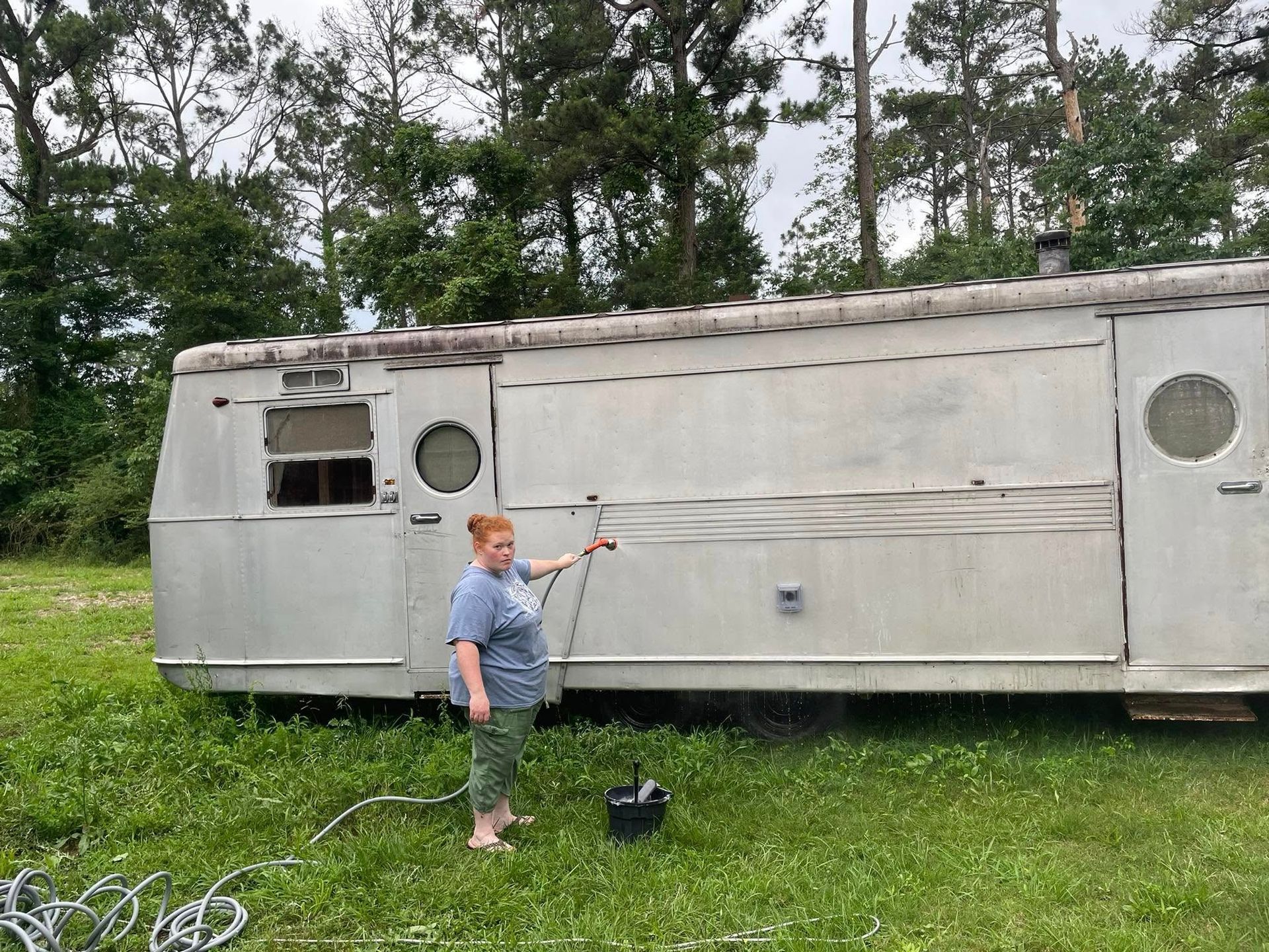 A person stands in a grassy field holding a hose against the side of a vintage silver trailer.