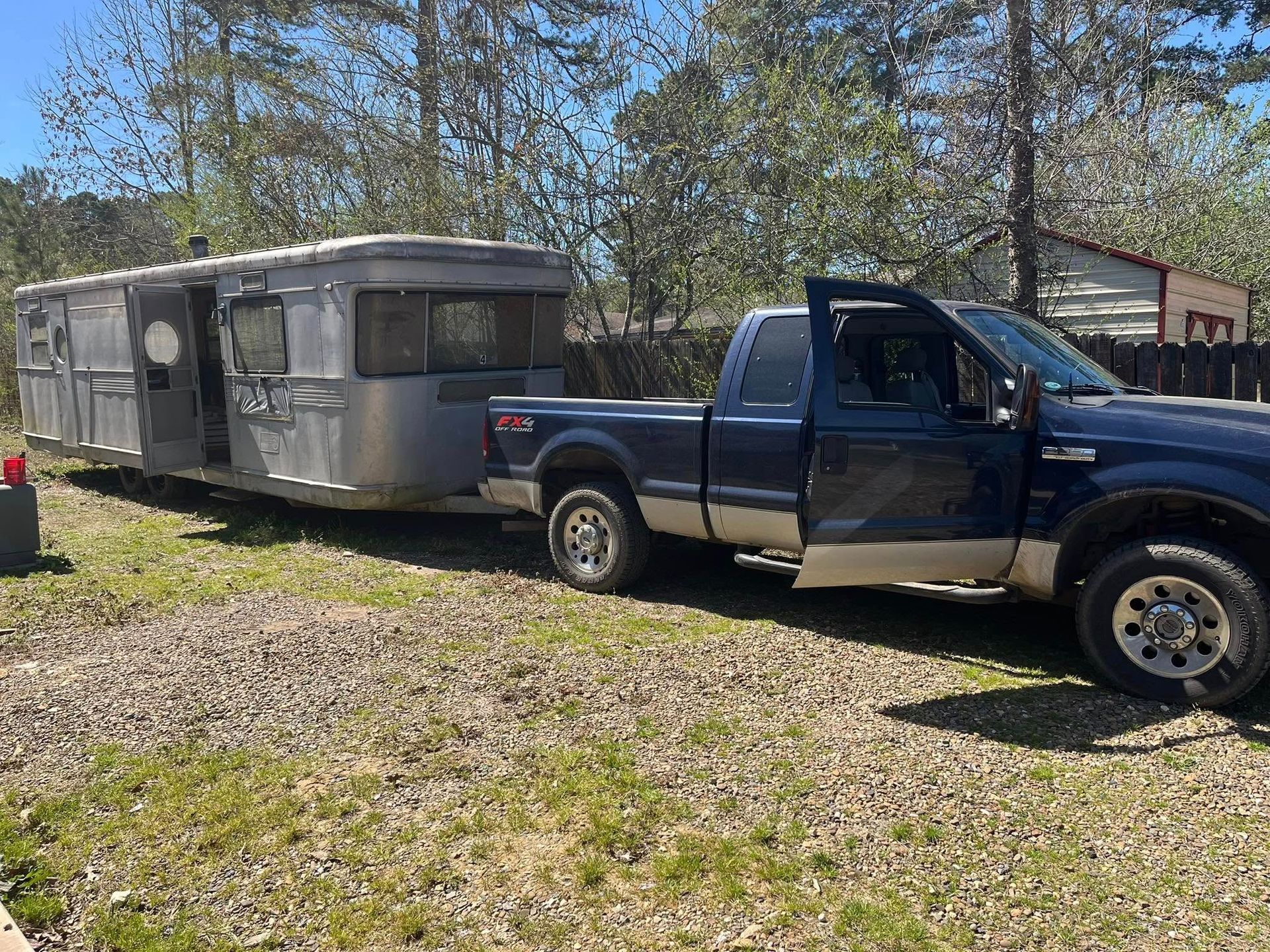 A dark blue Ford truck is parked on a gravel lot, towing a vintage silver travel trailer under a bright blue sky.