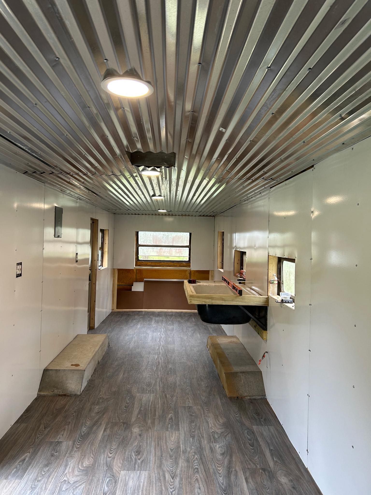 An empty trailer interior with white walls, a corrugated metal ceiling, wood-look flooring, and a small built-in counter.