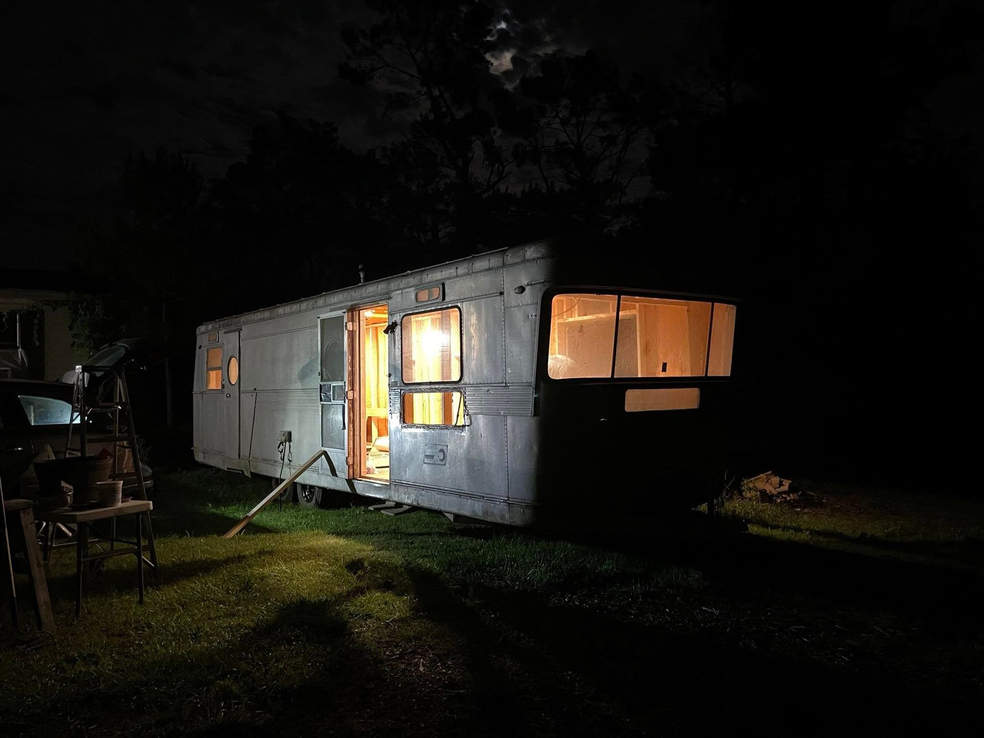 A weathered, boxy vintage camper trailer sits on a grassy field at night, glowing with warm light from its open door.