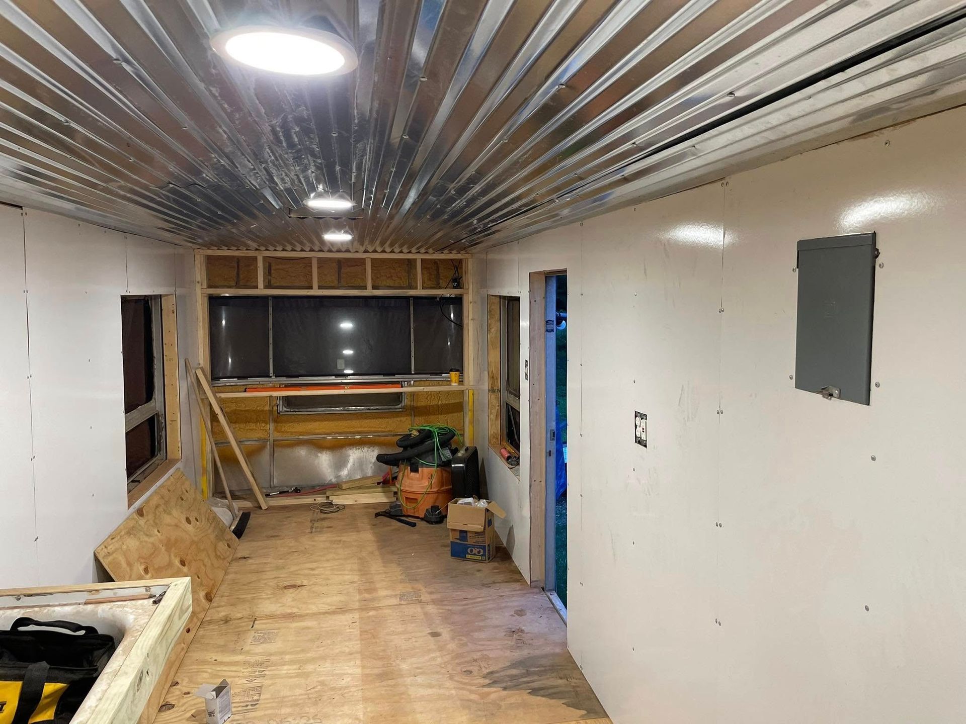Interior of a renovated trailer with corrugated metal ceiling, white wall panels, wooden flooring, and electrical panel.