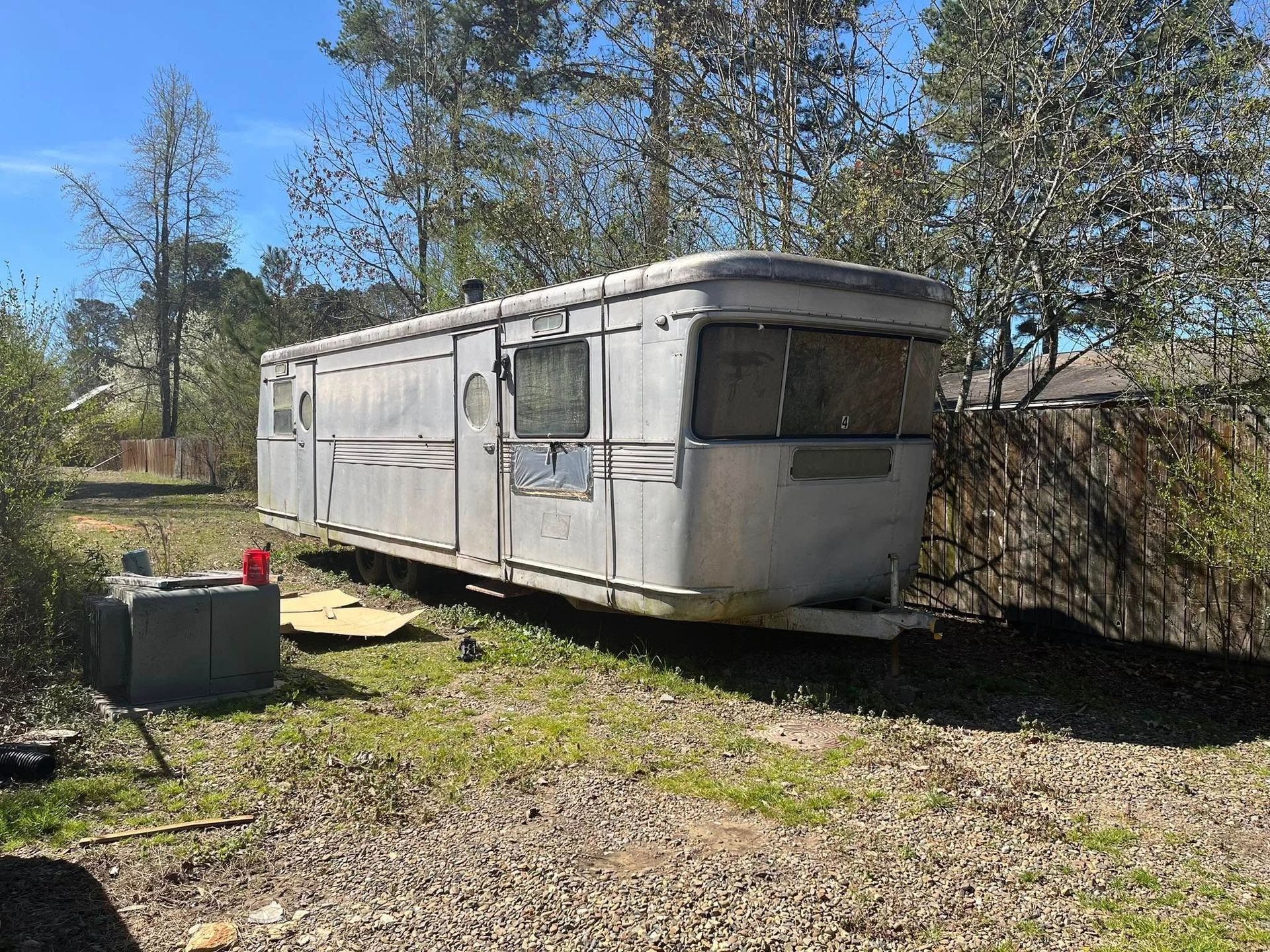 A weathered, silver vintage travel trailer parked on a grassy lot surrounded by trees on a sunny day.