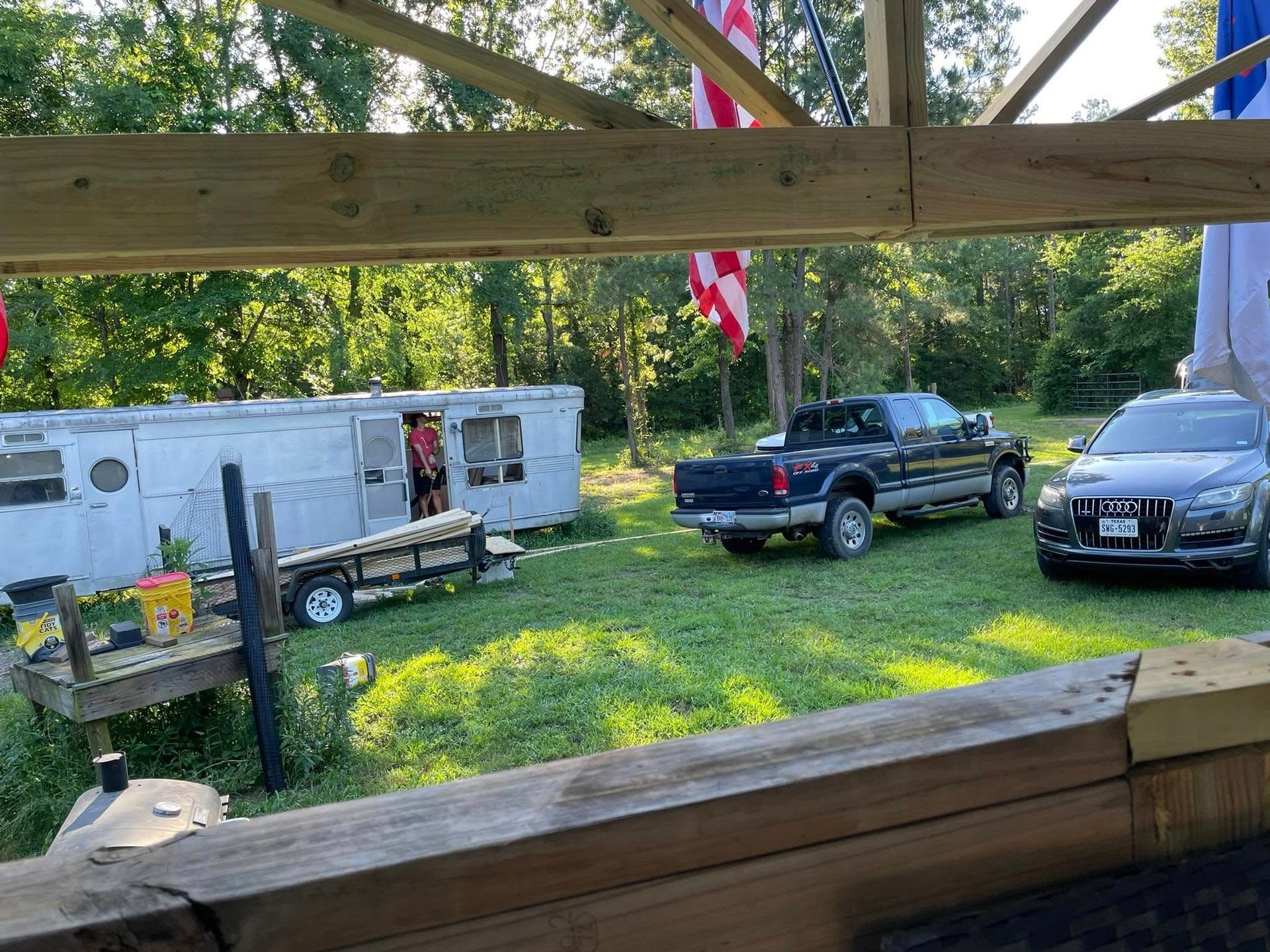 A view from under a wooden shelter shows a trailer, a dark pickup truck, and a grey SUV parked on a grassy lot with trees.