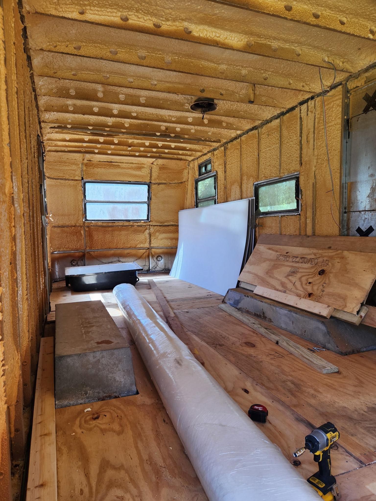 Interior of an unfinished camper trailer with foam-insulated walls, plywood floors, windows, and construction materials.
