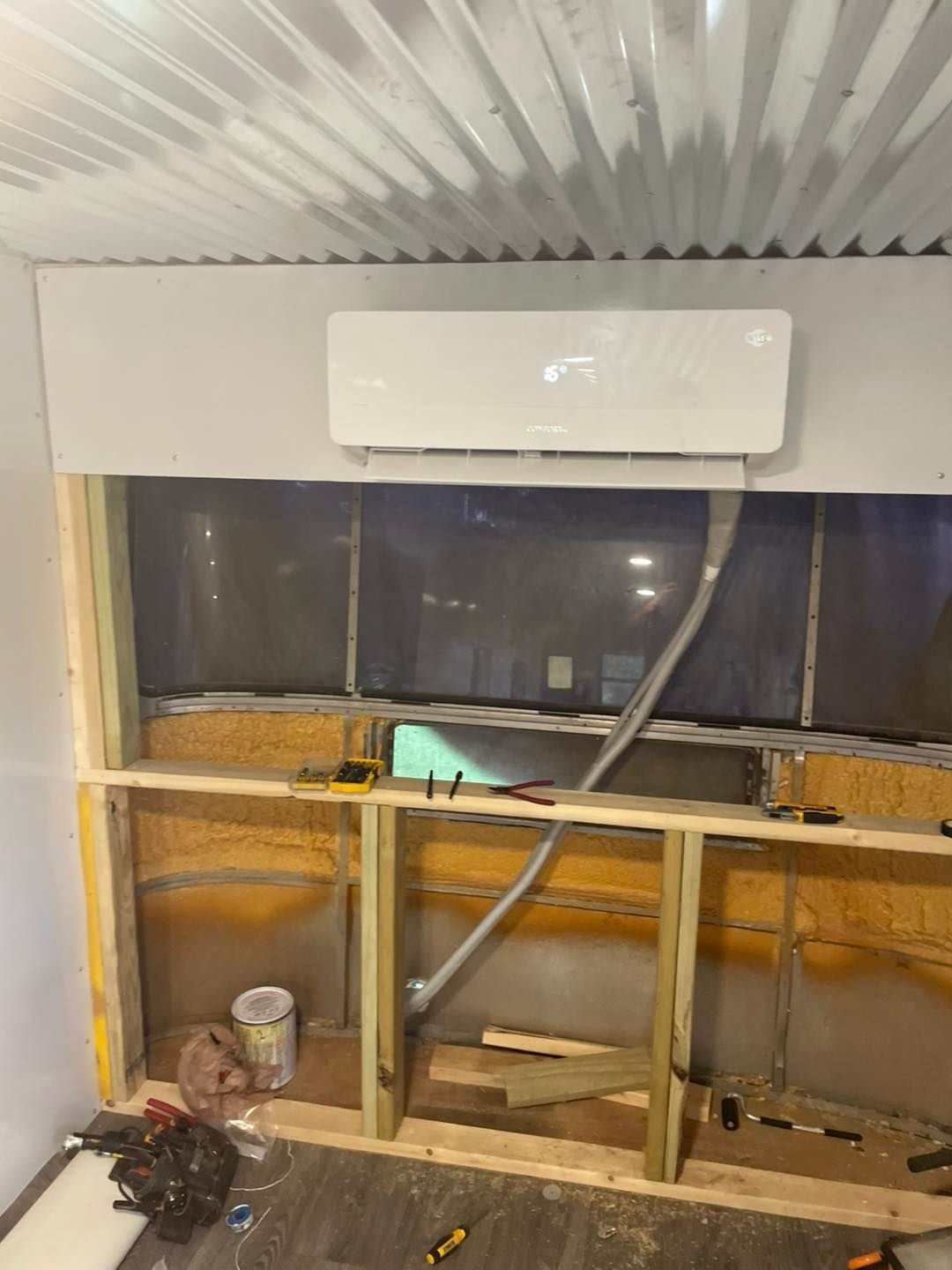 A wall-mounted air conditioner installed above a partially constructed wooden frame and corrugated ceiling in a room.