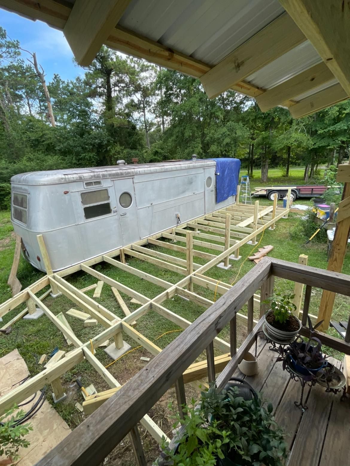 A vintage silver trailer sits next to a large, newly constructed wooden deck frame under construction in a grassy yard.