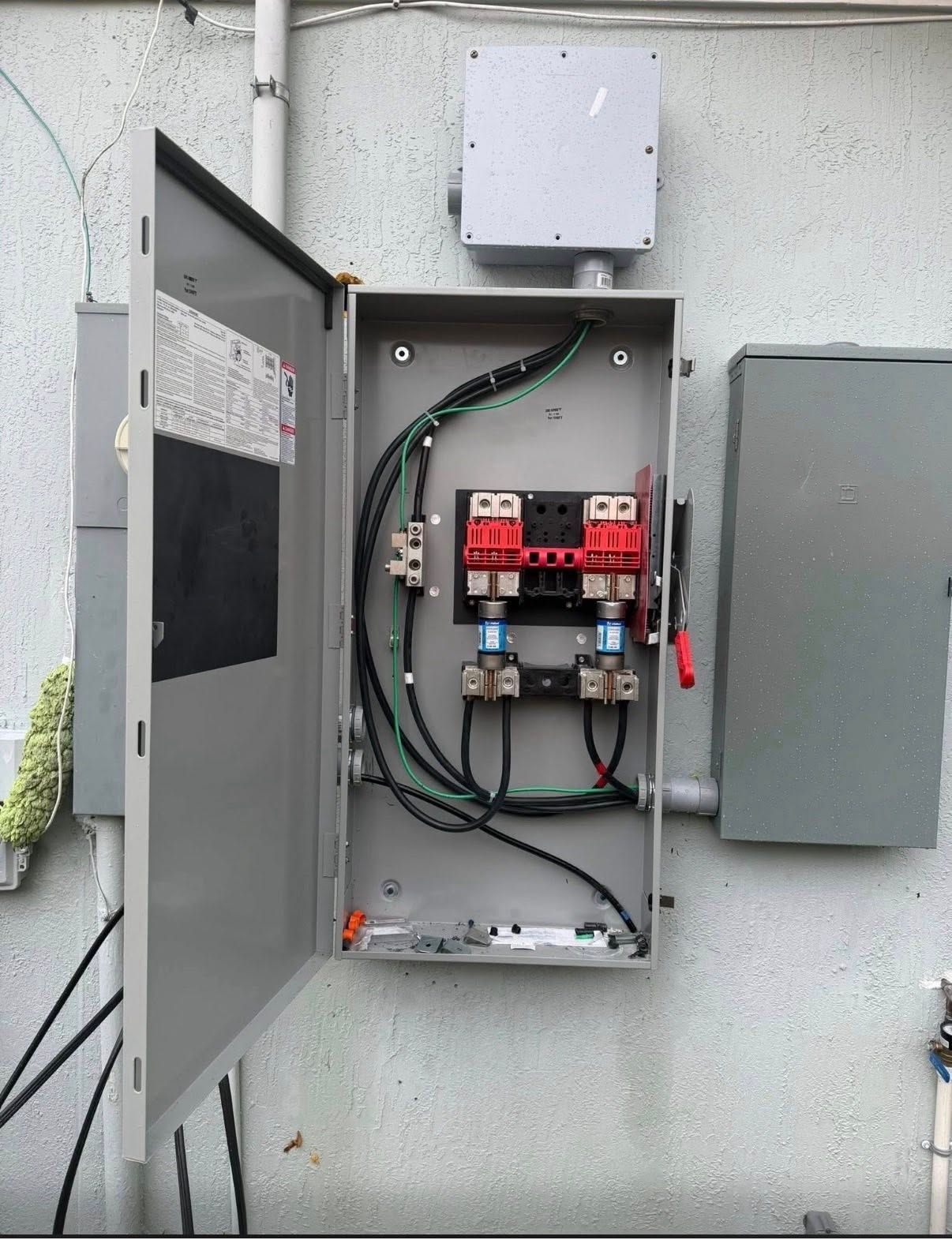 Open electrical breaker panel on an exterior wall with wires and adjacent gray box
