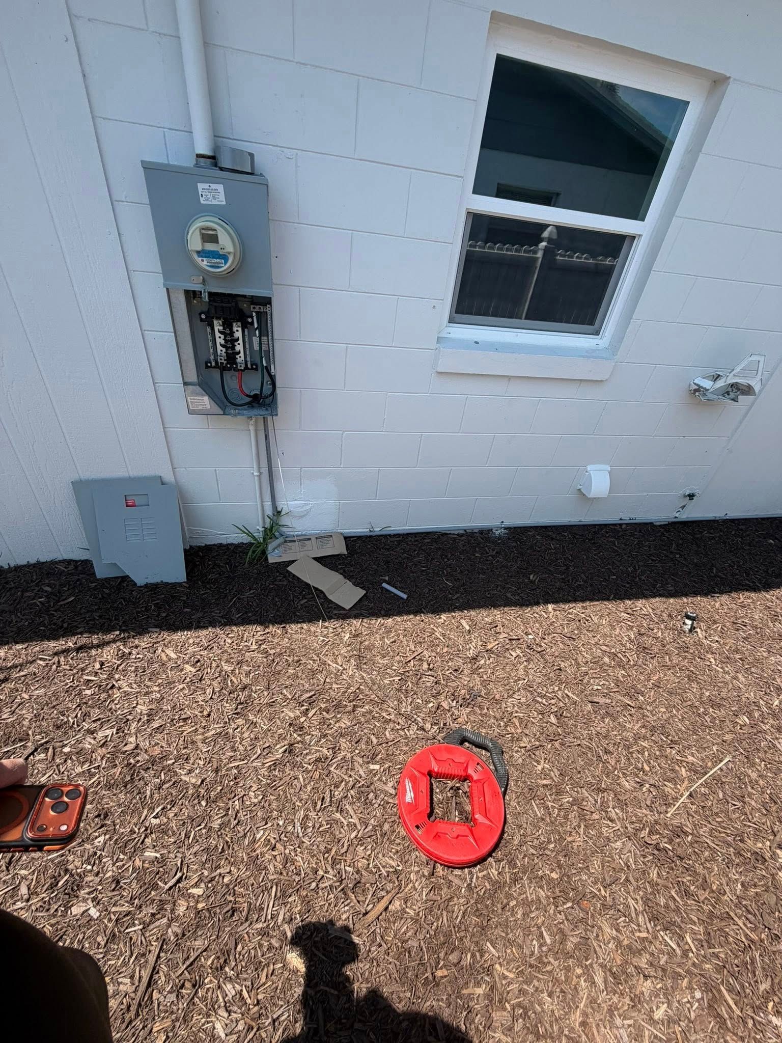 Exterior wall with an electrical meter, gray utility box, and a window above gravel ground.