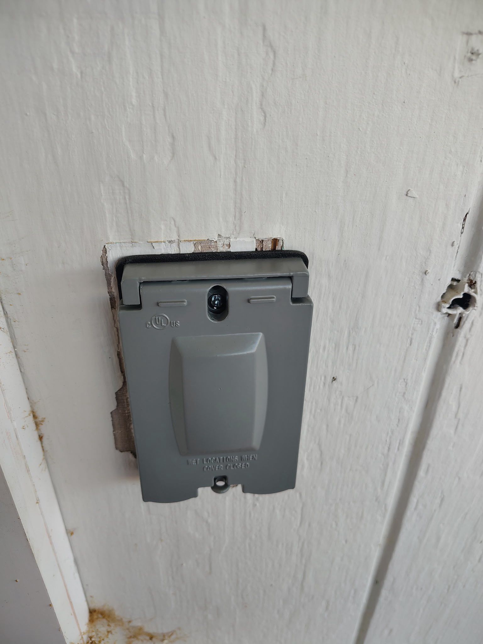Gray wall-mounted electrical outlet cover on a white wall with exposed trim edges.