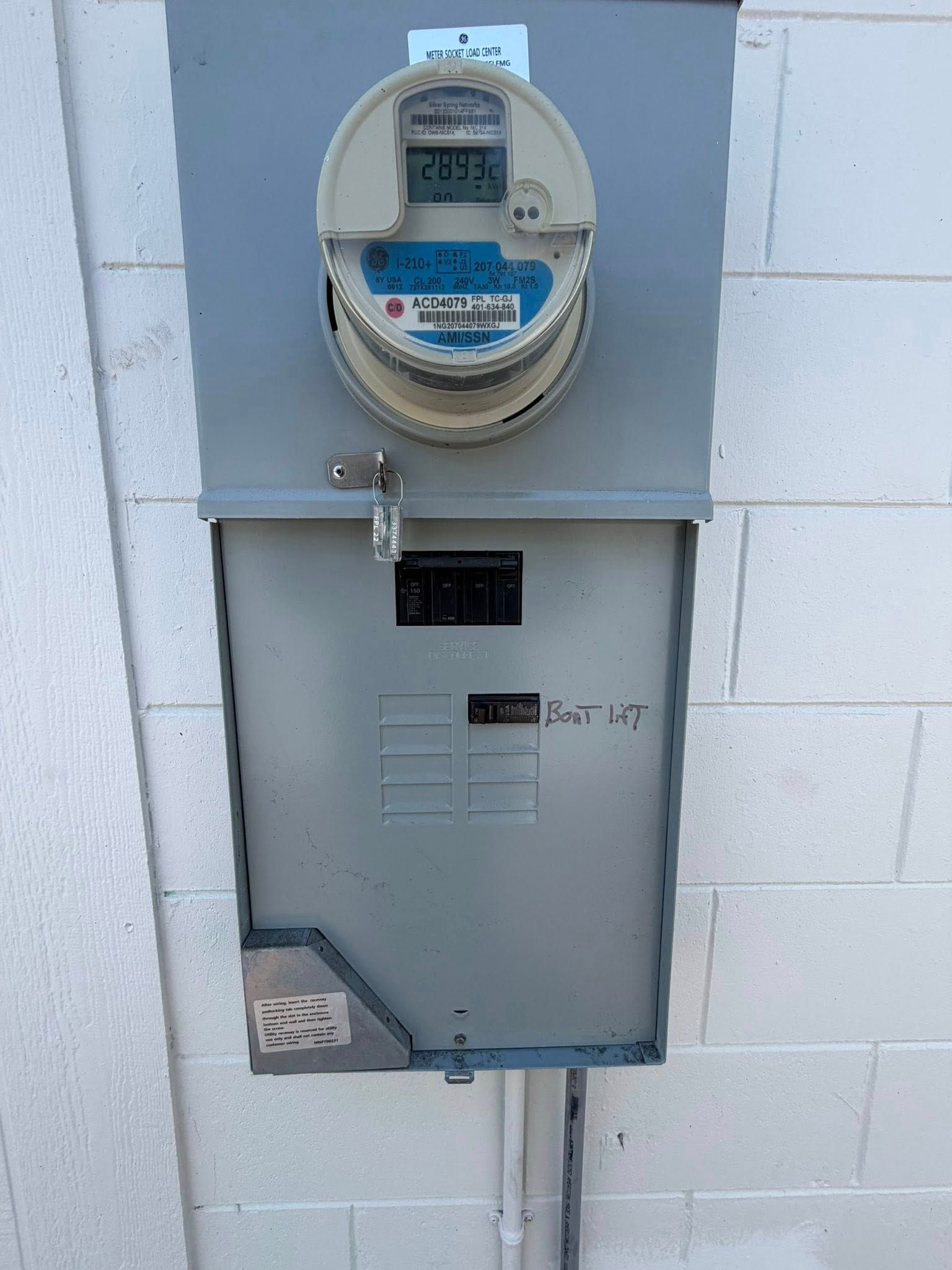 Gray outdoor electric meter box mounted on a white wall with conduit below.