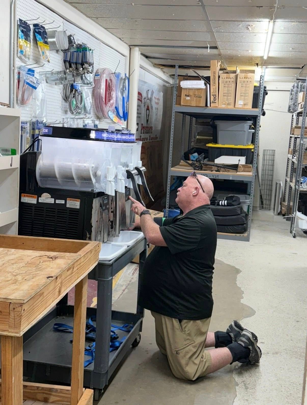 Kneeling man reaches toward supplies on a wall in a storage room lined with shelves and boxes.