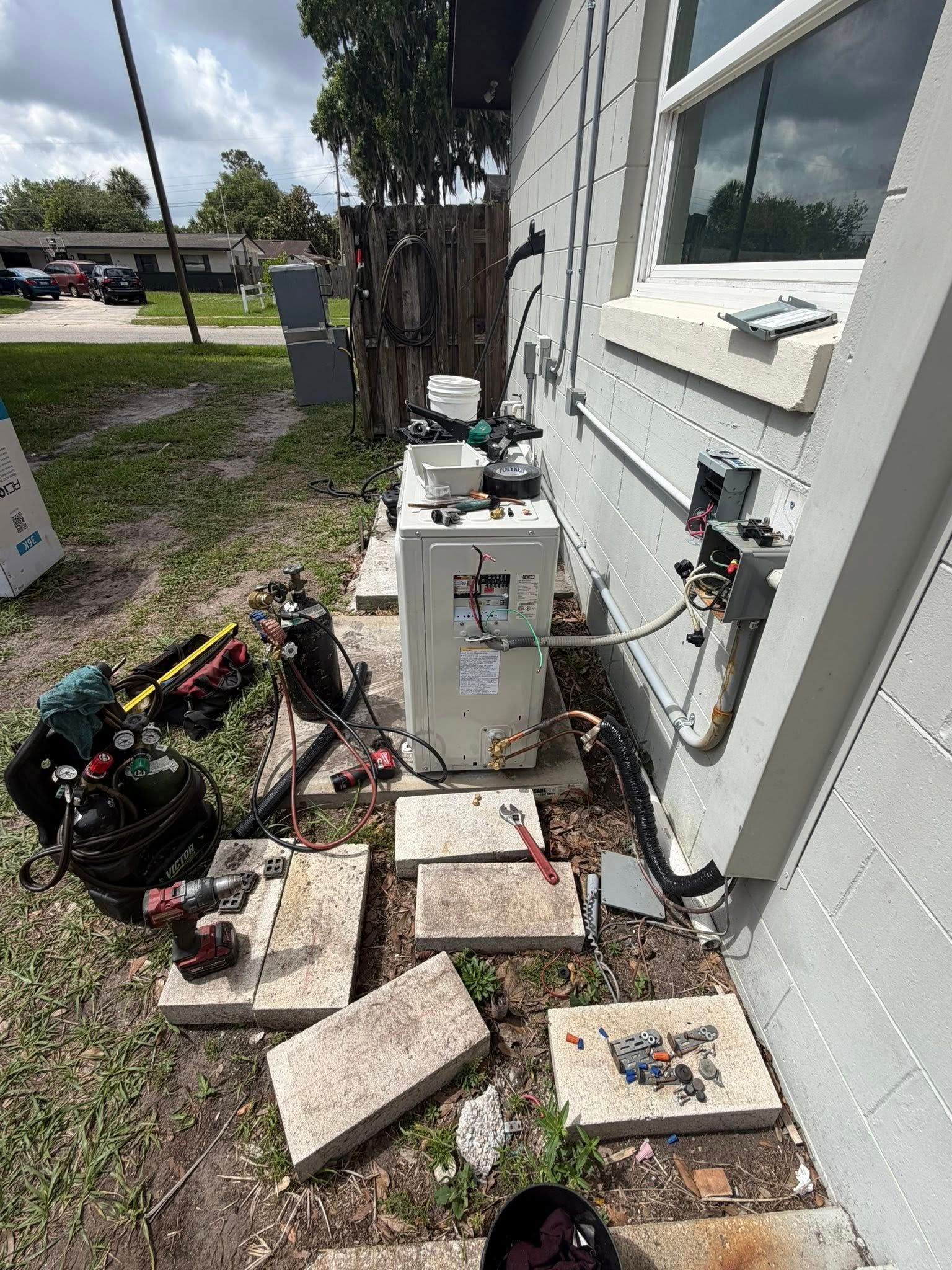 Outdoor utility area beside a house with HVAC equipment, scattered pavers, and tools on the ground.