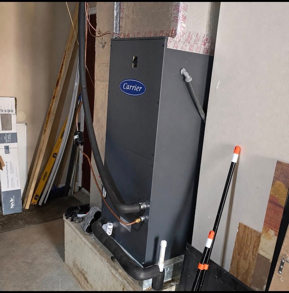 Gray HVAC/air handler unit in a utility room, with pipes and tools leaning nearby.