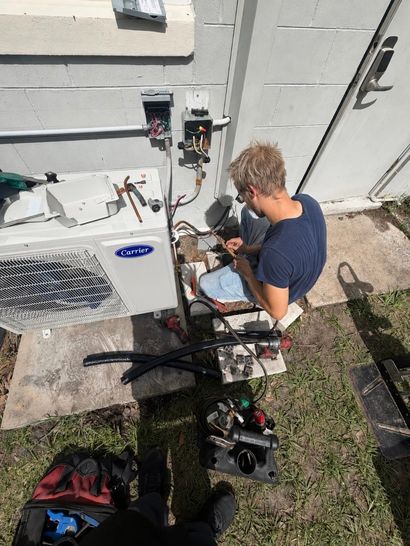 Person kneels by outdoor AC unit, checking wiring beside a building wall.
