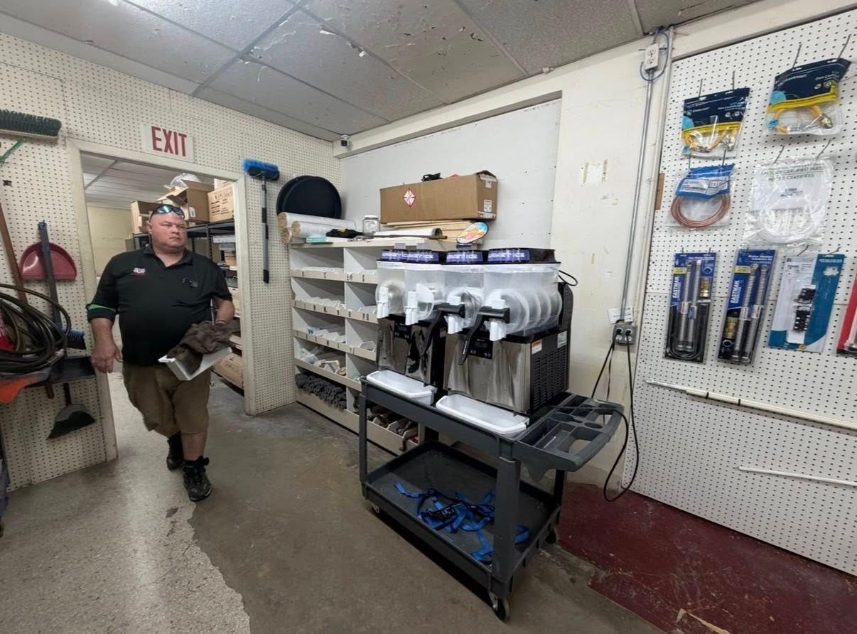 Man sitting in a utility room beside stacked boxes and shelving with industrial equipment and tools.