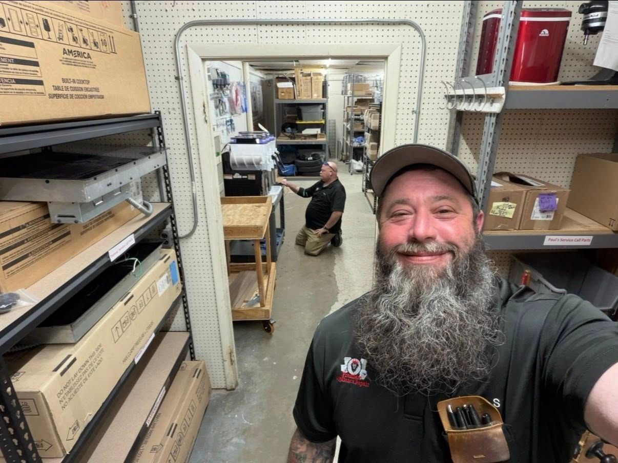 Man smiling in a workshop doorway, with shelves and a kneeling worker in the background.