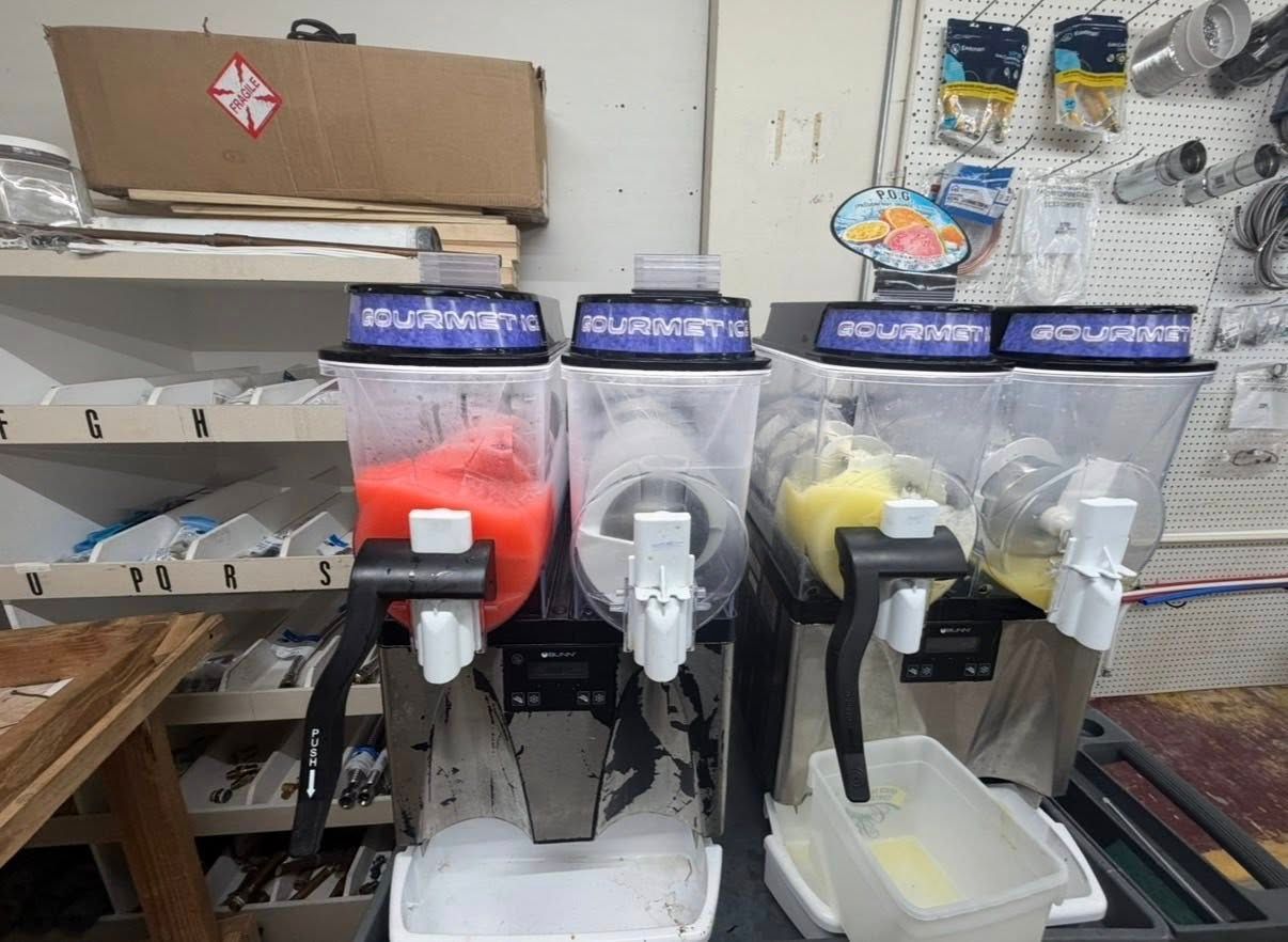 Four beverage dispensers with colored liquids lined up on a counter in a kitchen or café setting