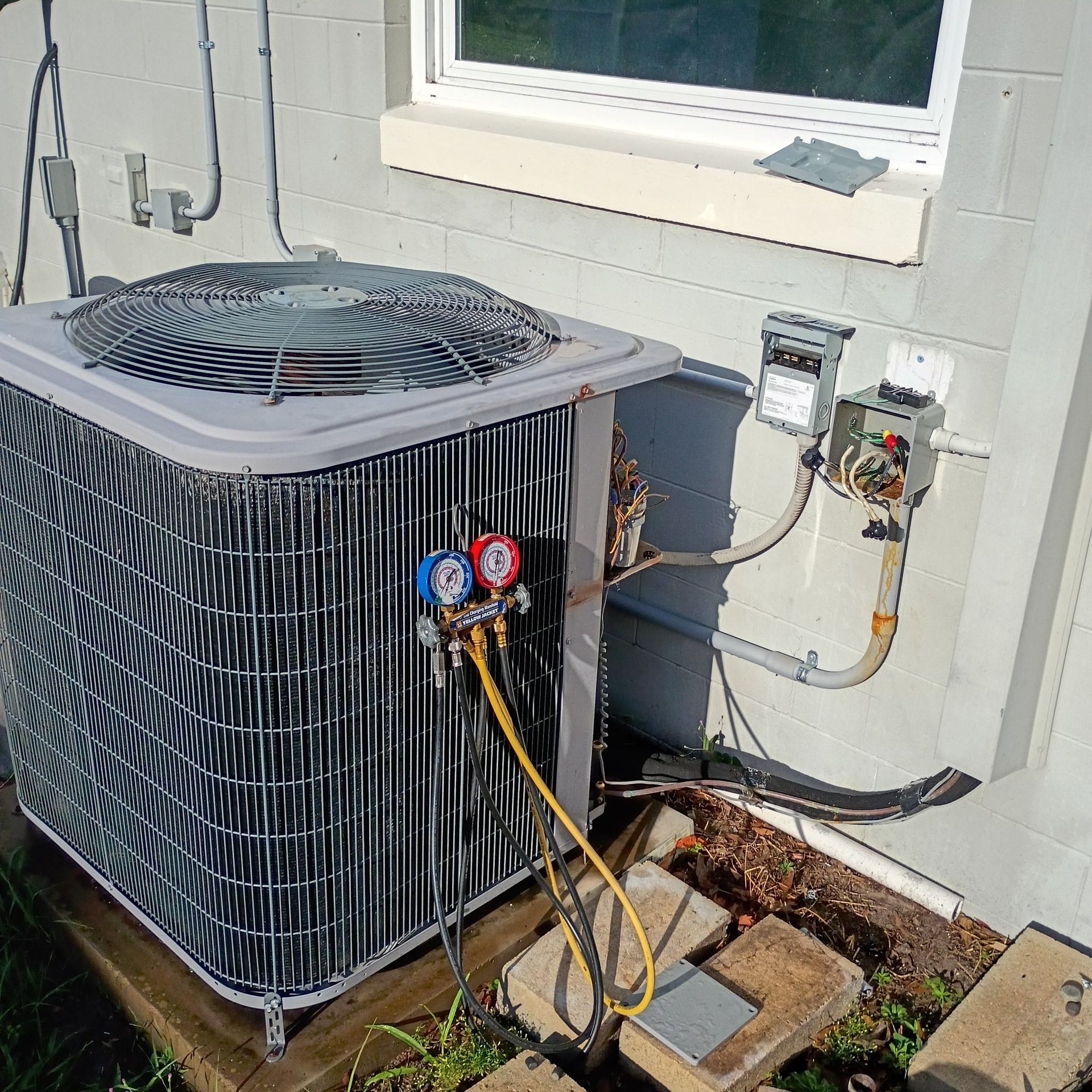 Outdoor air conditioner unit beside house wall with gauges and yellow hoses attached, under a window.