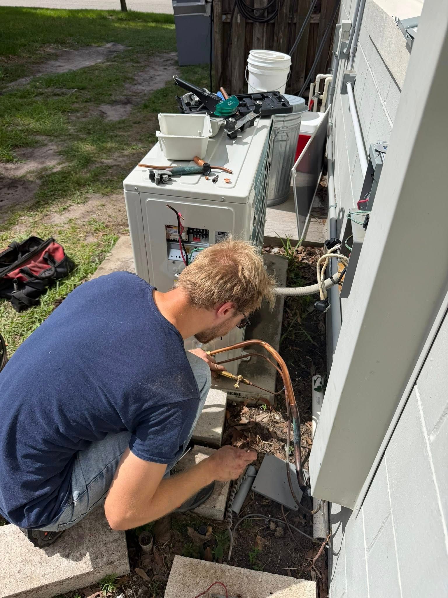Person crouches beside an outdoor HVAC unit, using a tool to repair wiring.