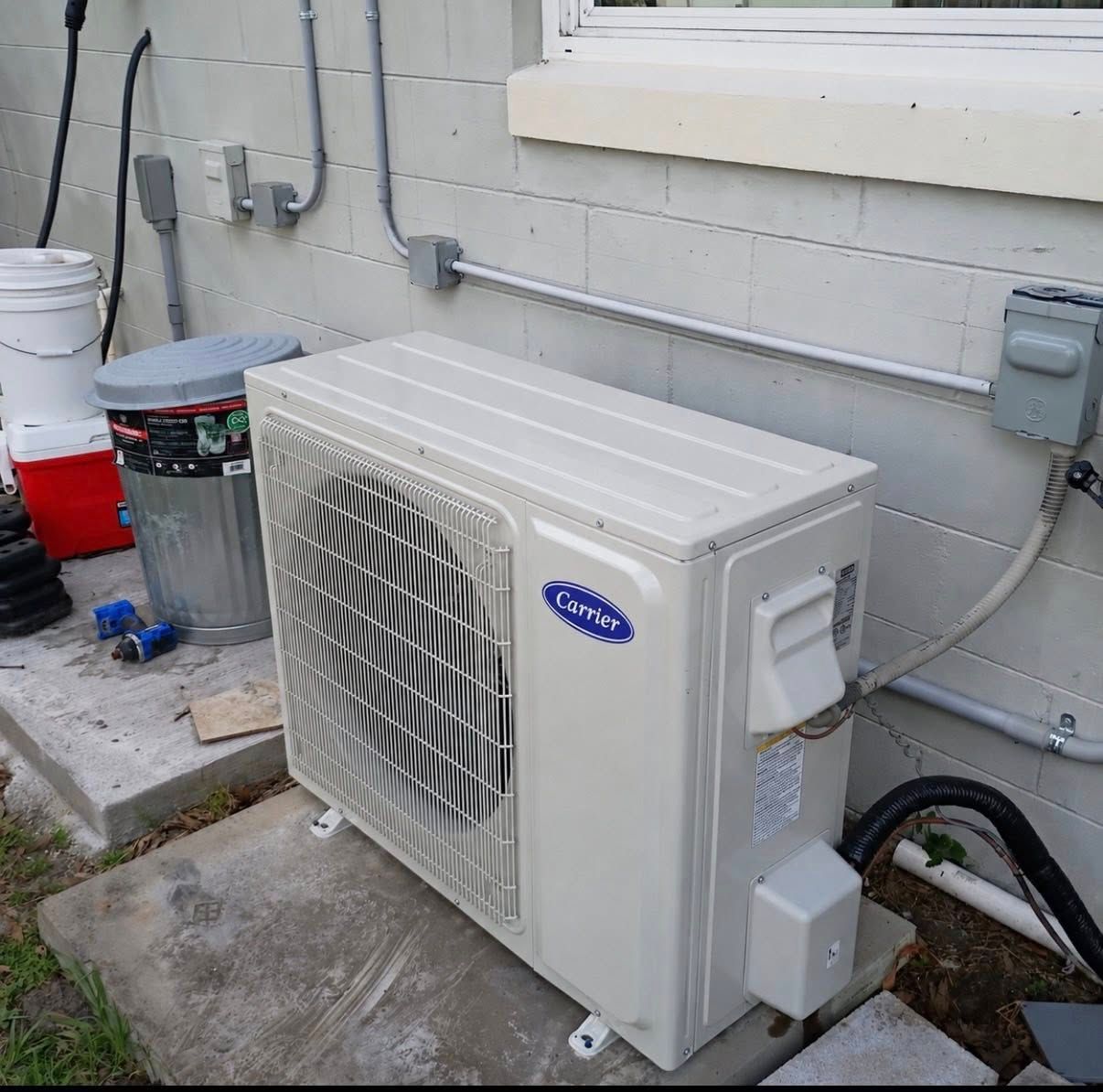 White outdoor air conditioner unit beside a house wall with pipes and buckets on a concrete slab