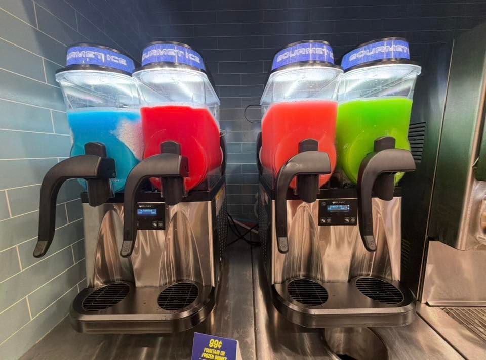 Four frozen drink dispensers in blue, red, orange, and green at a stainless steel counter.