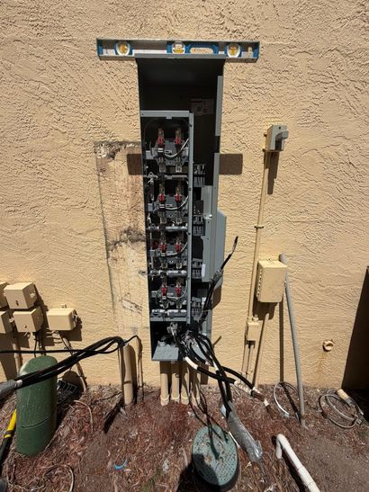 Open electrical panel in stucco wall with exposed wiring and a hanging keypad on the right.