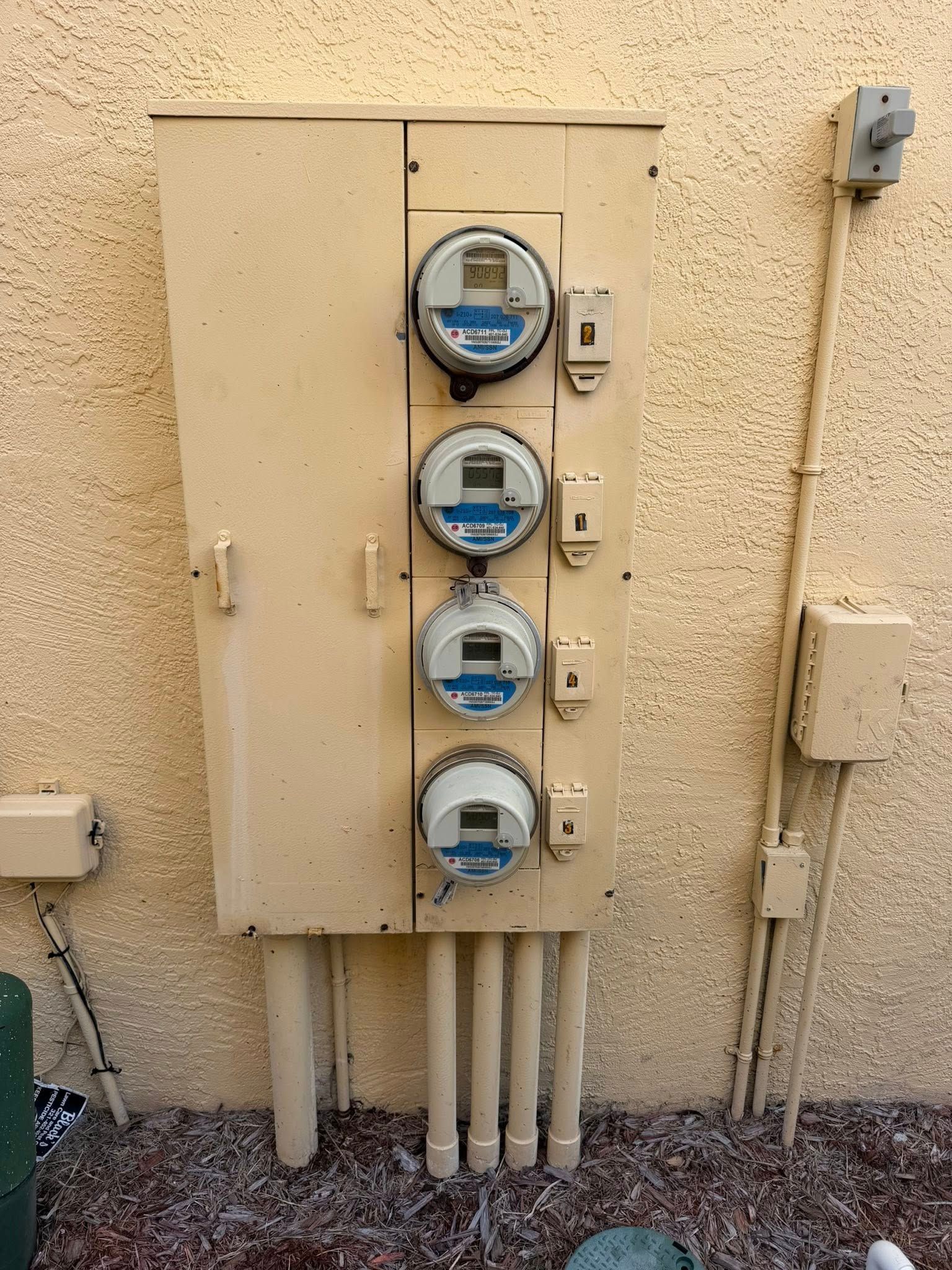 Four electric meters on a beige utility panel mounted on a stucco exterior wall.
