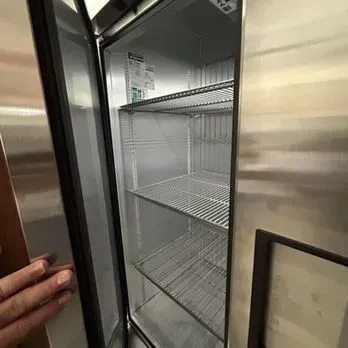 Open stainless-steel refrigerator with empty wire shelves being held open by a hand