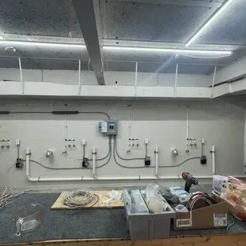 Industrial utility room with wall-mounted pipes, valves, and wiring above a cluttered workbench.