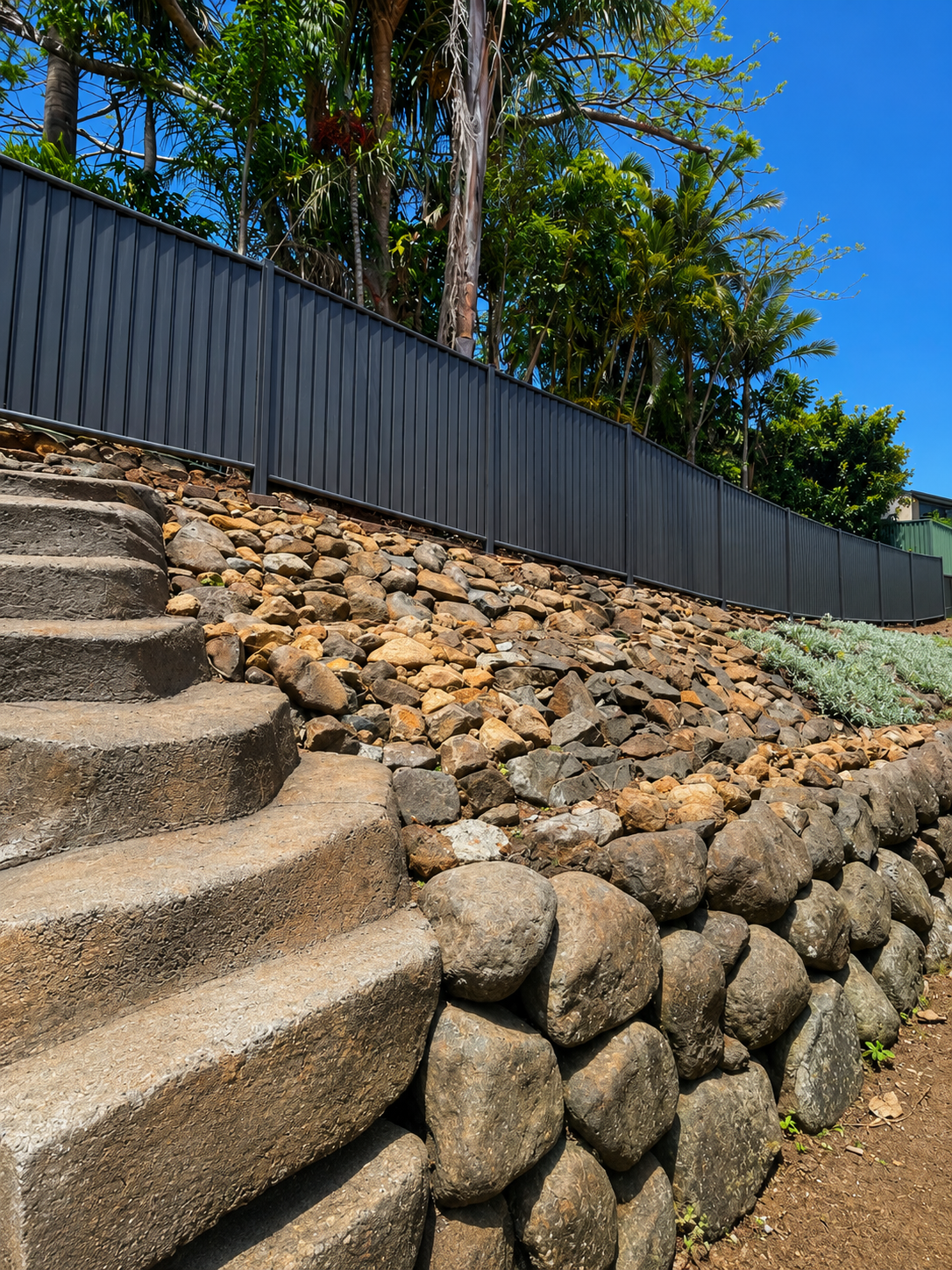 Stepped stone retaining wall with grey fence and trees along a sunny hillside path