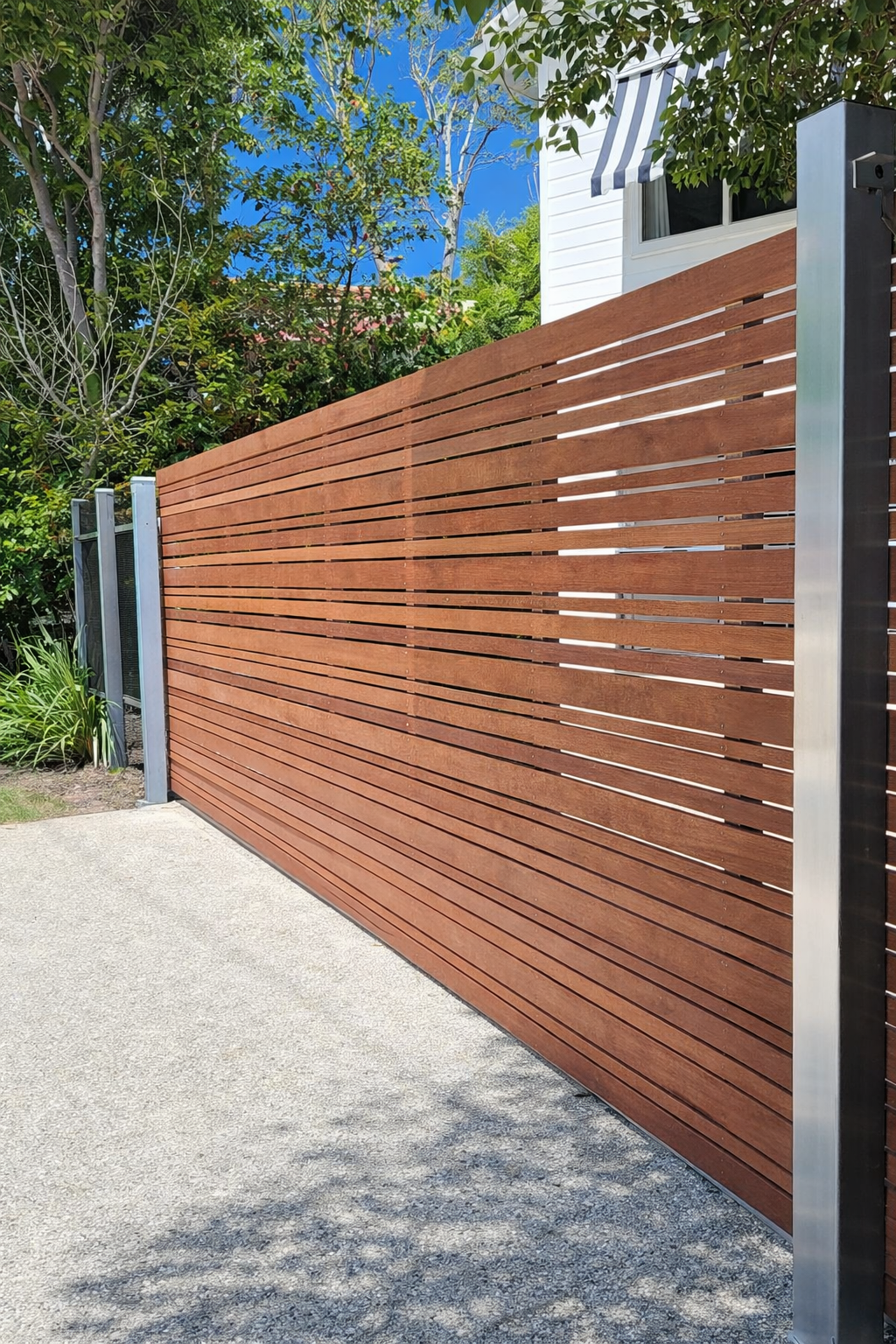 Modern wooden slat fence with grey metal posts beside a driveway and trees