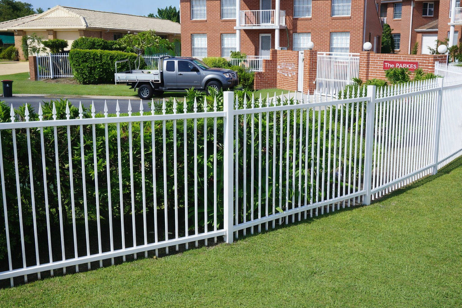 White metal fence along a green lawn in front of a brick house and driveway with a parked pickup truck.