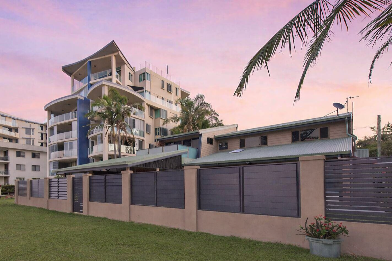 Modern apartment complex behind a low wall at sunset with a pink sky and palm trees