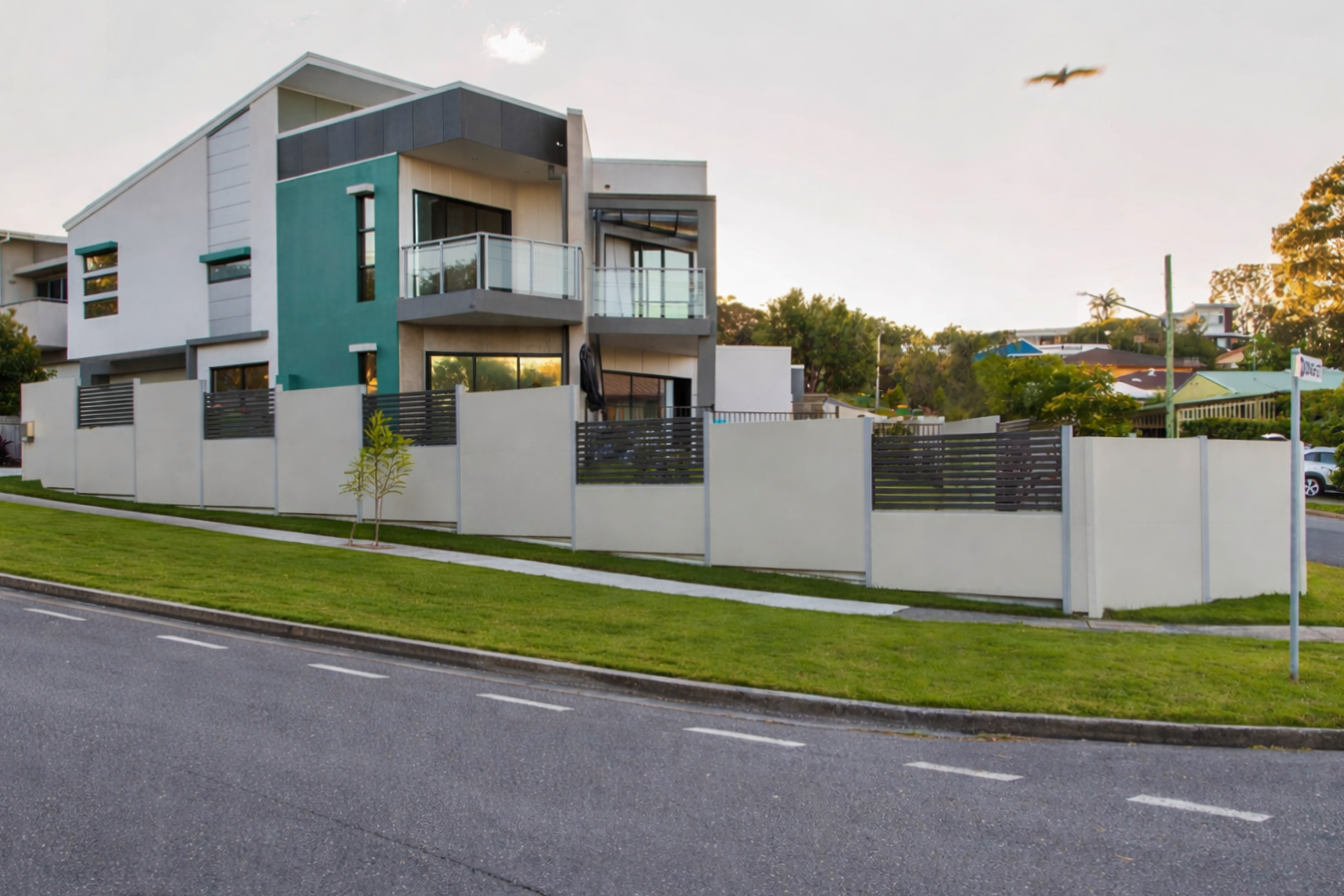 Modern two-story house with balconies, white fence, and landscaped street corner under a clear sky