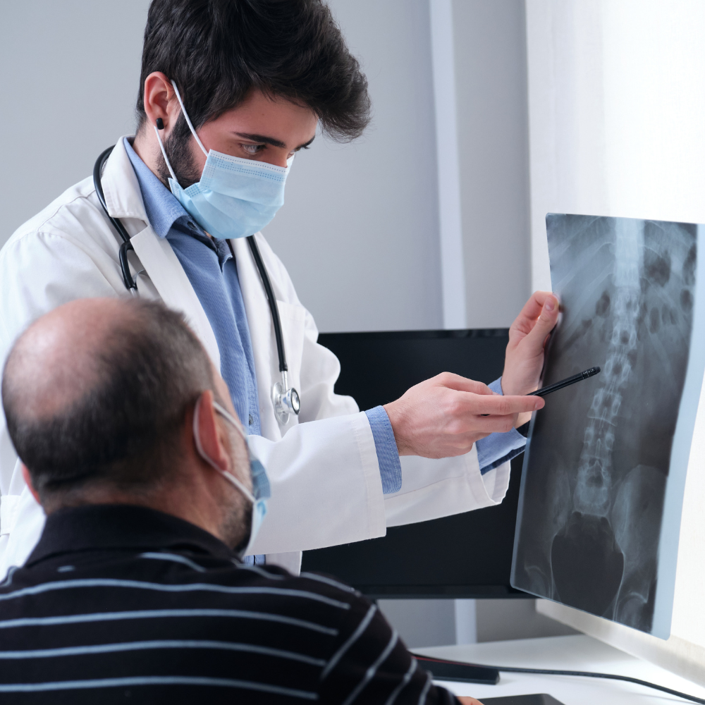 Doctor in mask pointing at spine X-ray, explaining to a patient.
