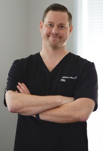 Man in black scrubs with arms crossed, smiling, in front of a window.