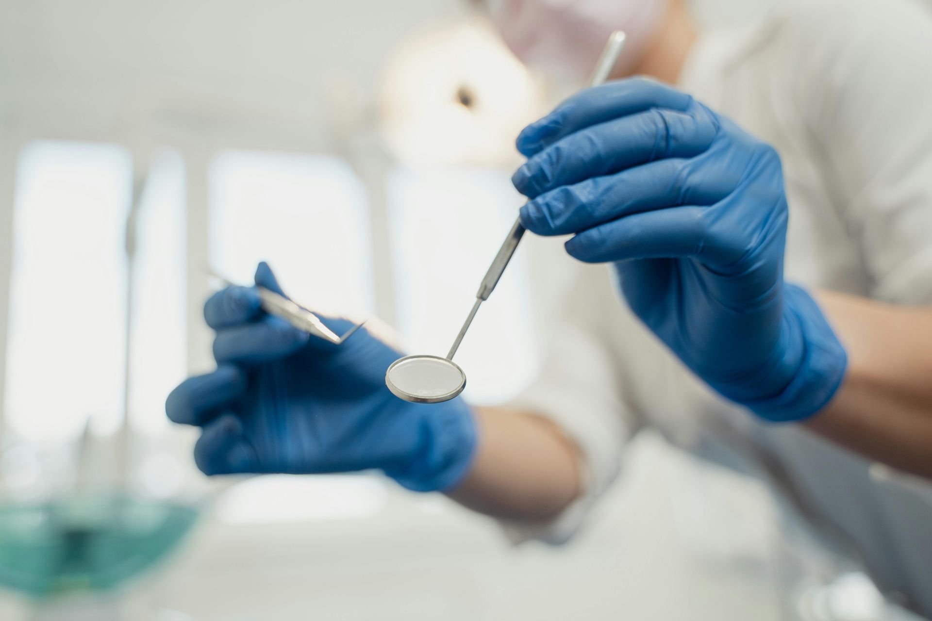 Dentist wearing blue gloves holding dental mirror and probe in a bright examination room.