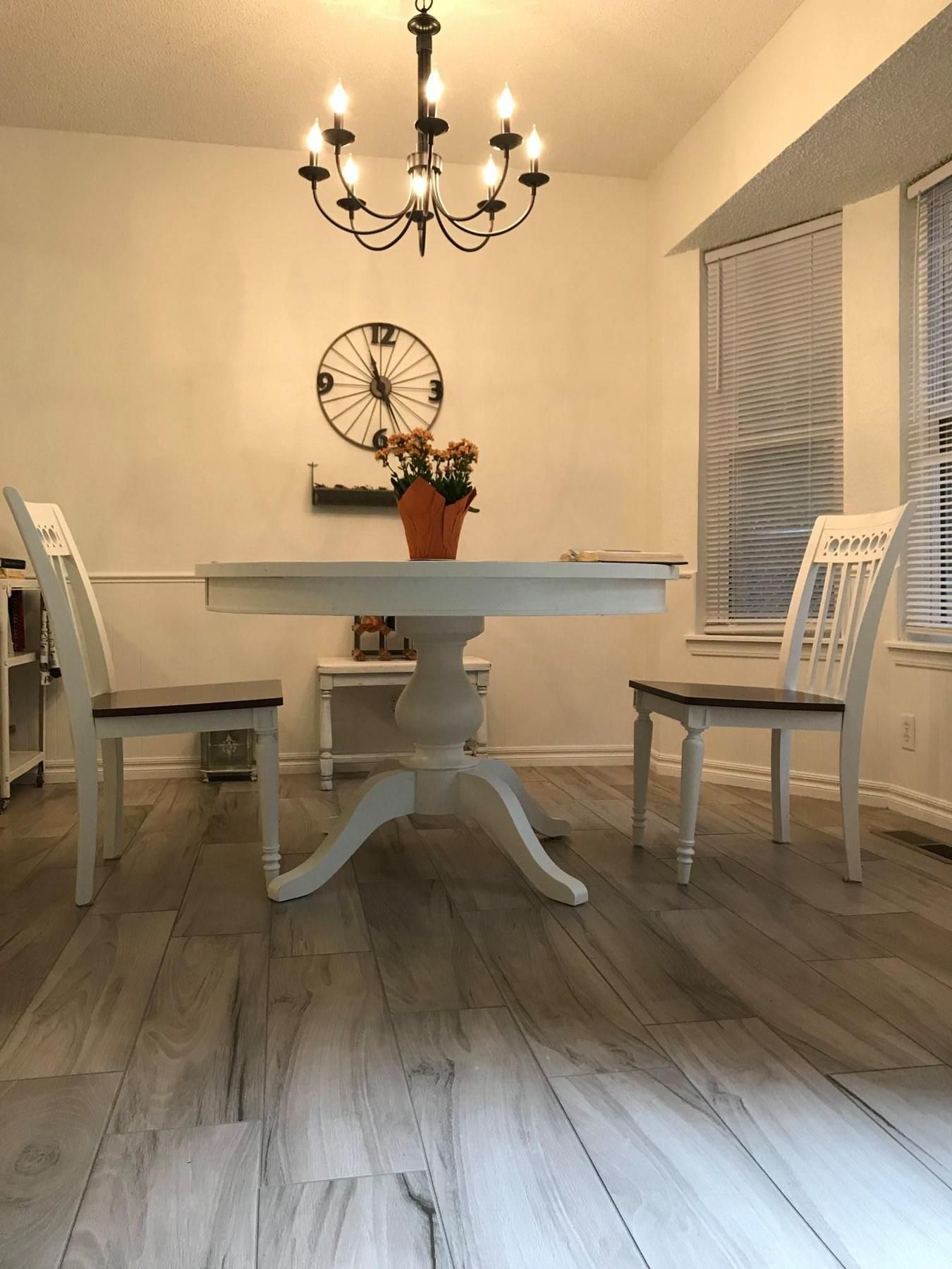 A bright dining room with a white pedestal table, two chairs, a clock, and a chandelier. Gray wood-look flooring.