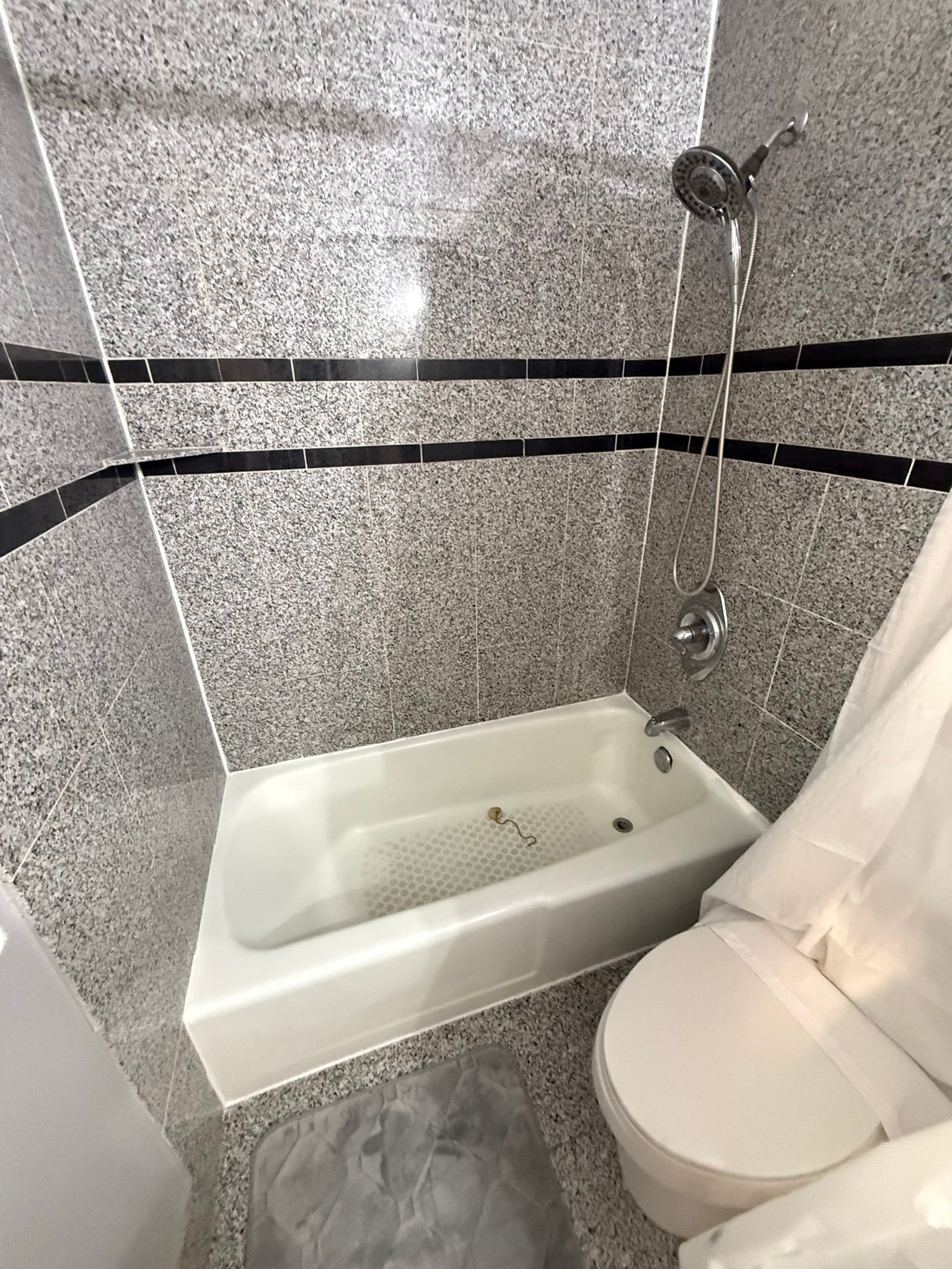 Small bathroom with tub, shower, toilet, and granite tile walls with black accent stripes.