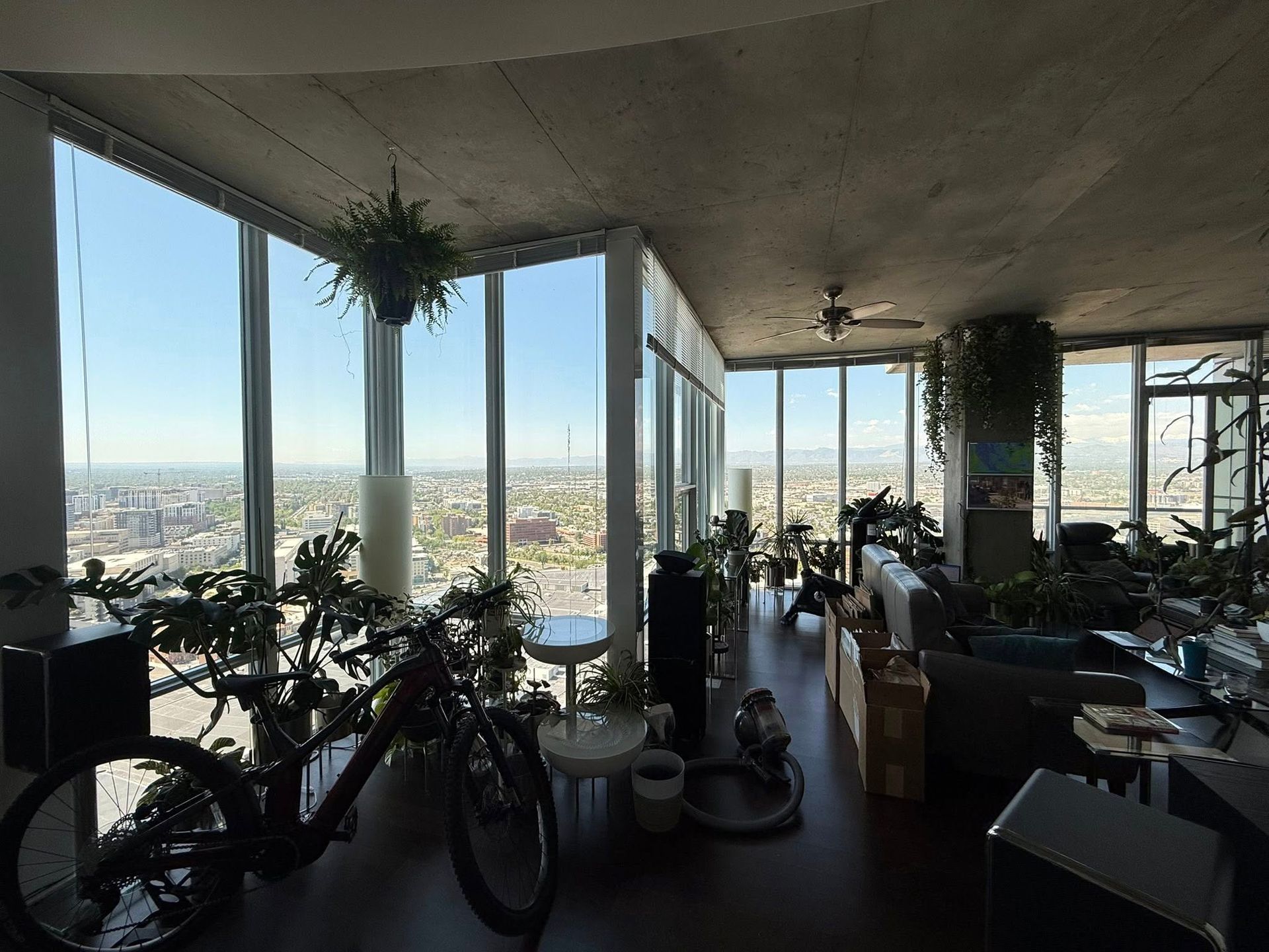 Modern living room with floor-to-ceiling windows, city view, plants, and a bicycle inside.