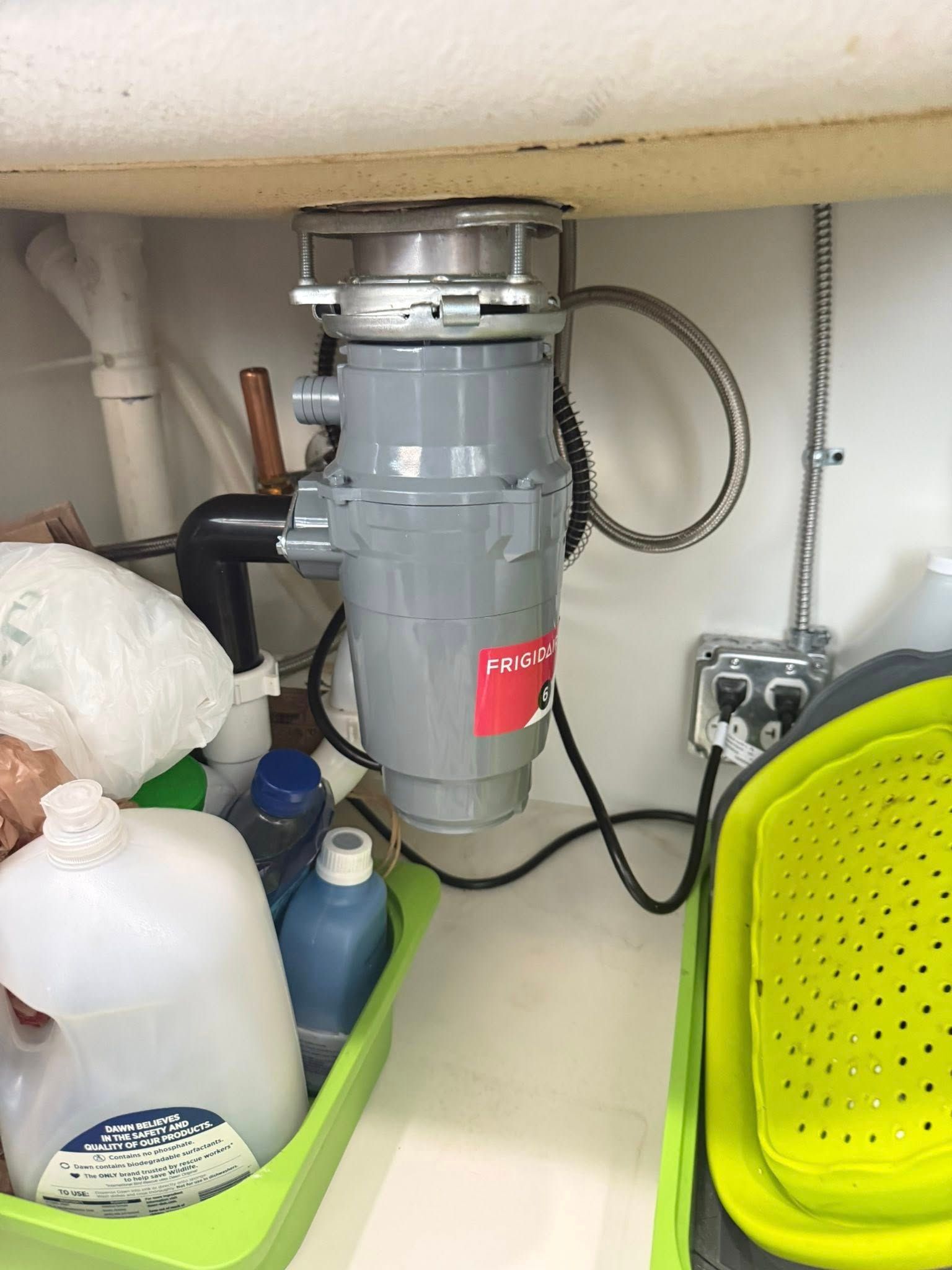 Garbage disposal unit under a kitchen sink. The unit is gray and attached to plumbing and an electrical outlet.