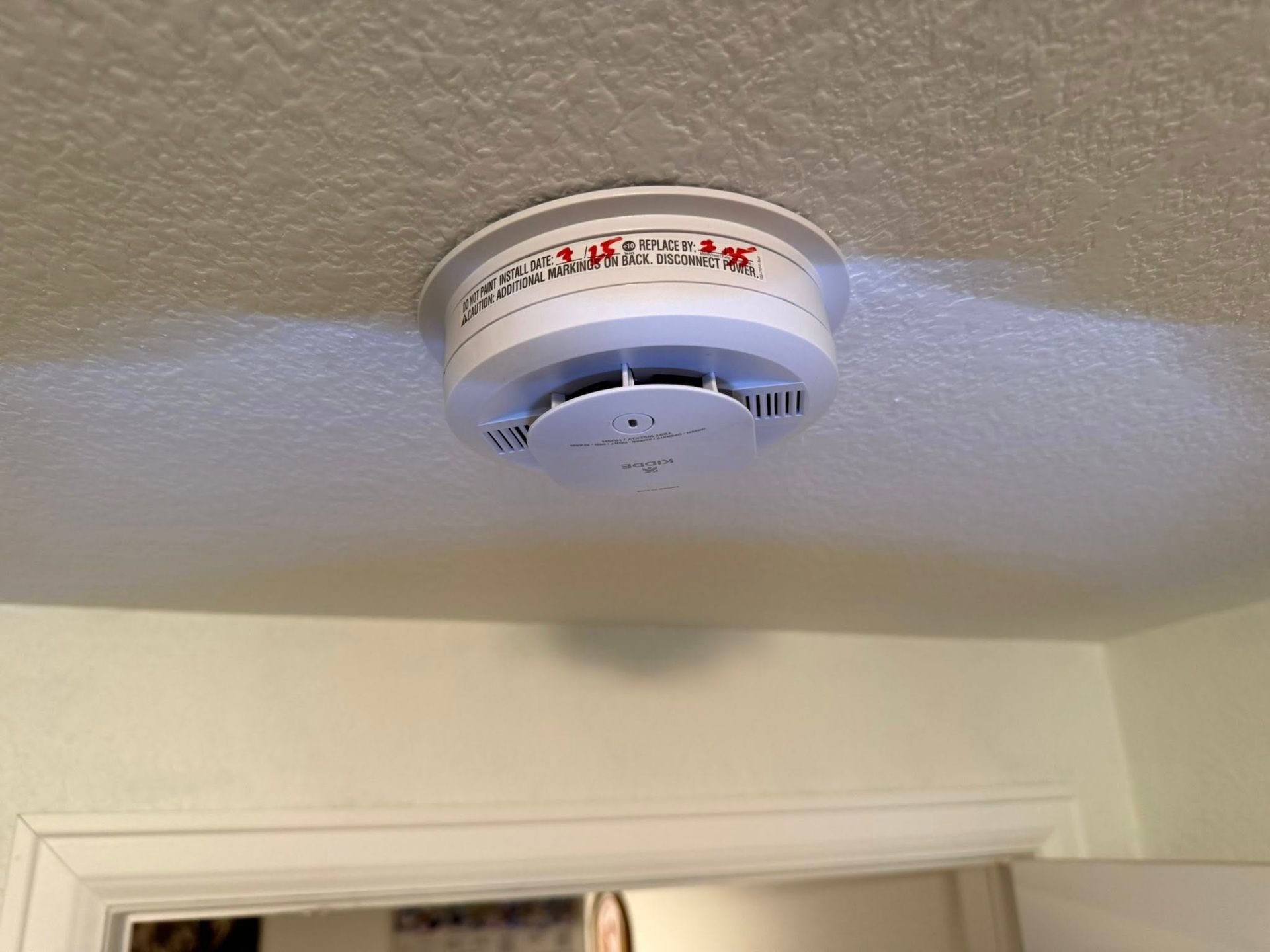 White smoke detector mounted on a white textured ceiling, above a doorway.