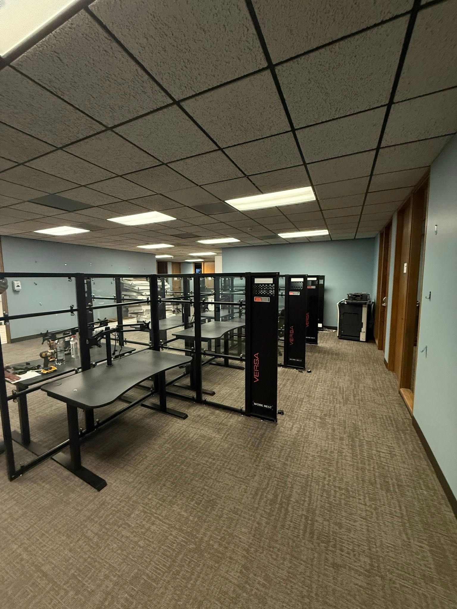 Office cubicles with gray desks and black dividers; carpeted floor and tile ceiling.