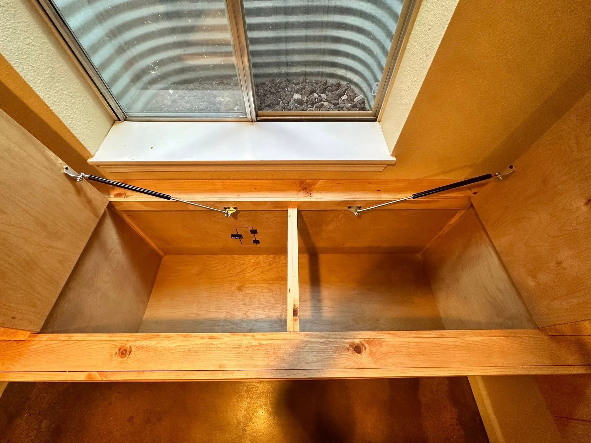 Wooden storage box built under a window, featuring a split interior and support hinges.