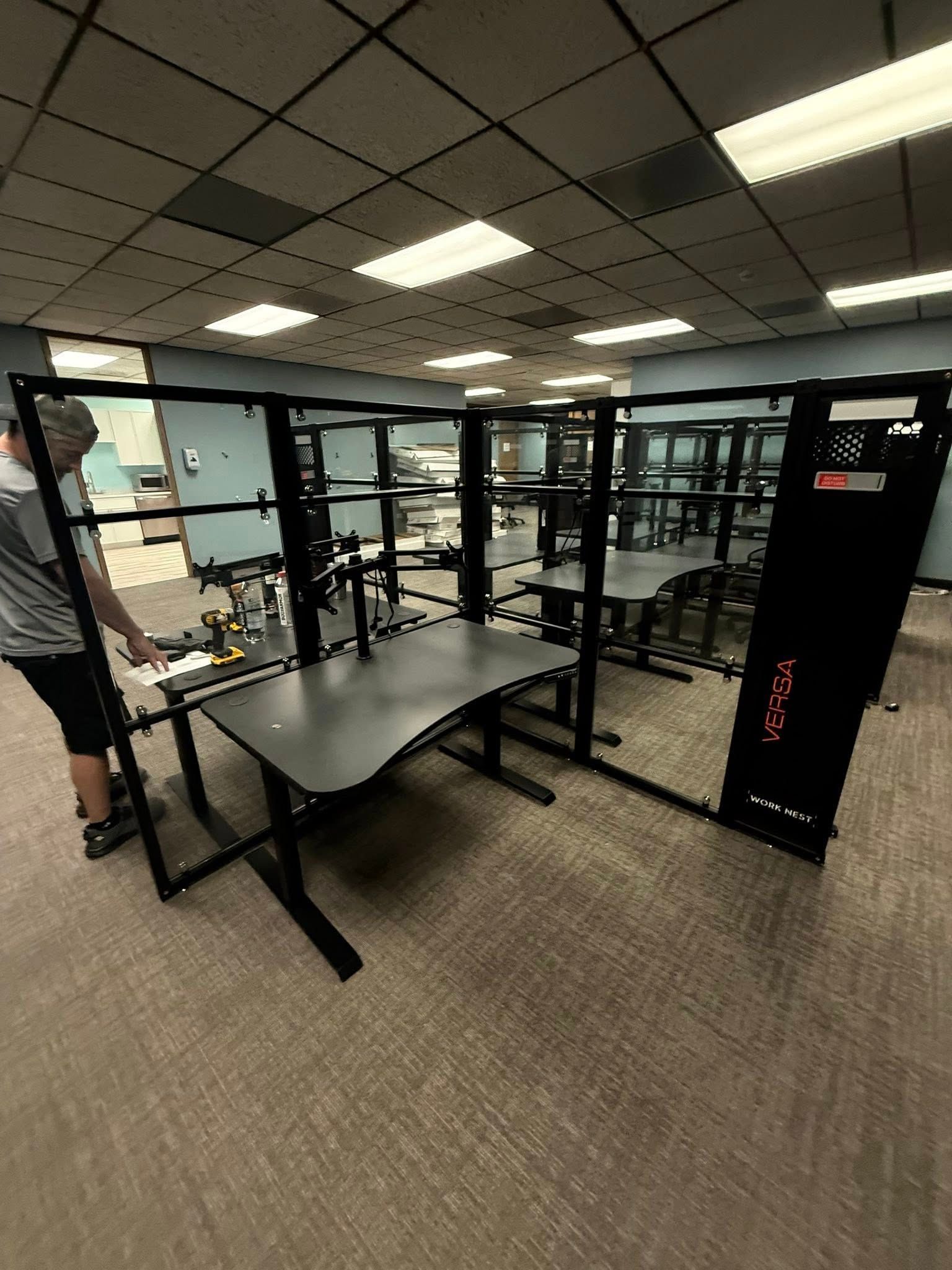Person setting up a black gaming desk within a black partitioned room. The room has overhead lighting and carpet.