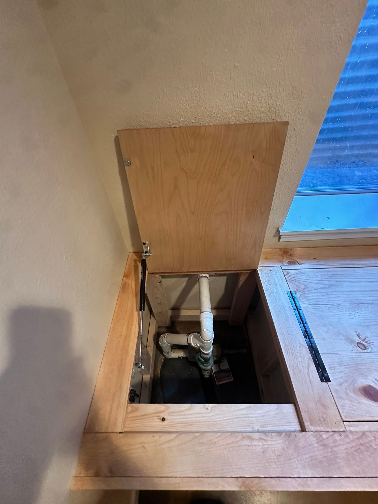 Plywood door open, revealing plumbing in a built-in cabinet under a slanted ceiling, near a window.