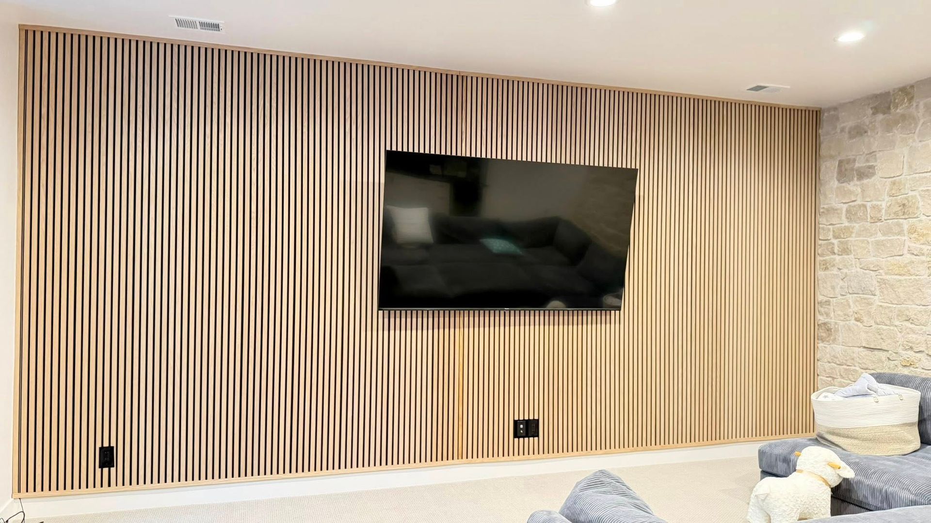 Wooden slatted wall with a mounted TV; room with stone accent wall and grey sofa.