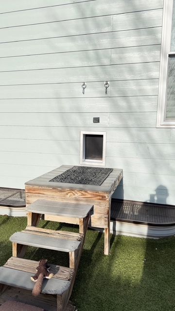 Wooden dog house with stairs and pet door against a light blue building; artificial turf.