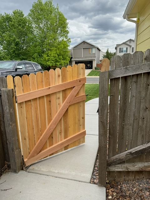 Wooden fence gate, open on a concrete path, leading to a street. House visible in the background.