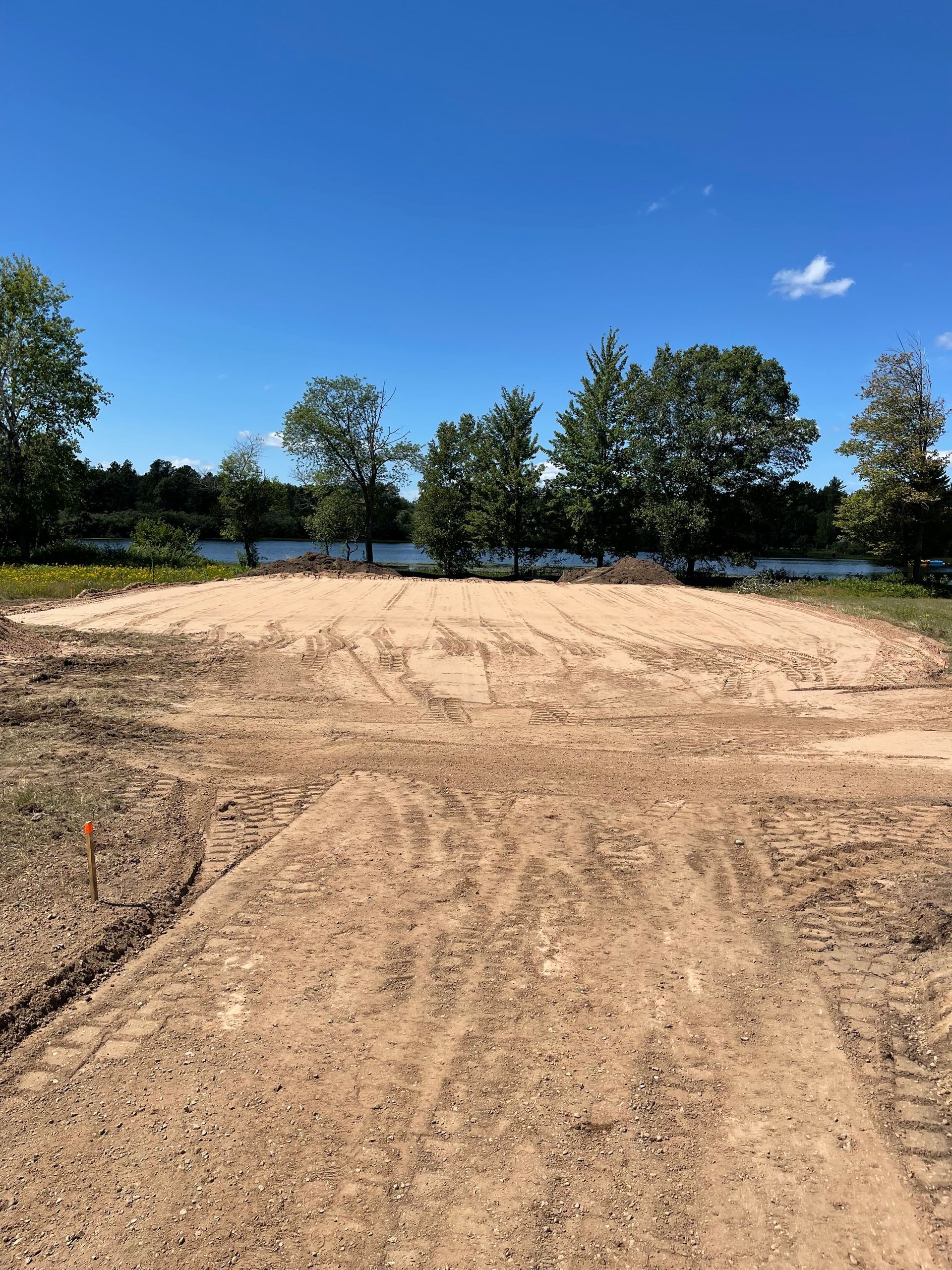 Dirt patch with tractor tracks, trees, and water under a blue sky.